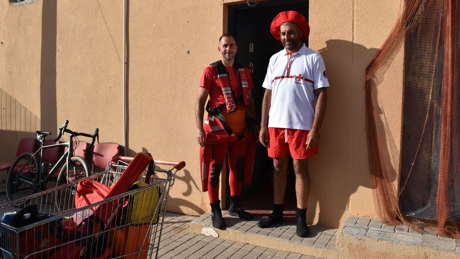 Los voluntarios de Cruz Roja José Antonio Carracao y Mohamed Chergui a las puertas de la base de la ONG en el puerto pesquero. En primer plano, a la izquierda, el carrito de la compra que usan para transportar el equipo necesario desde la base hasta el barco