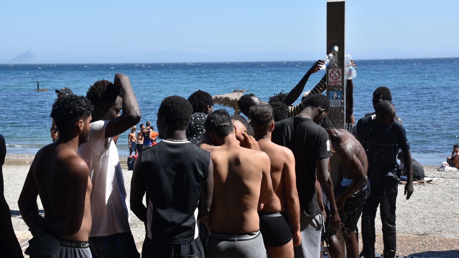 Una fila de personas migrantes para asearse y llenar botellas de agua en las duchas de la playa del Trampolín
