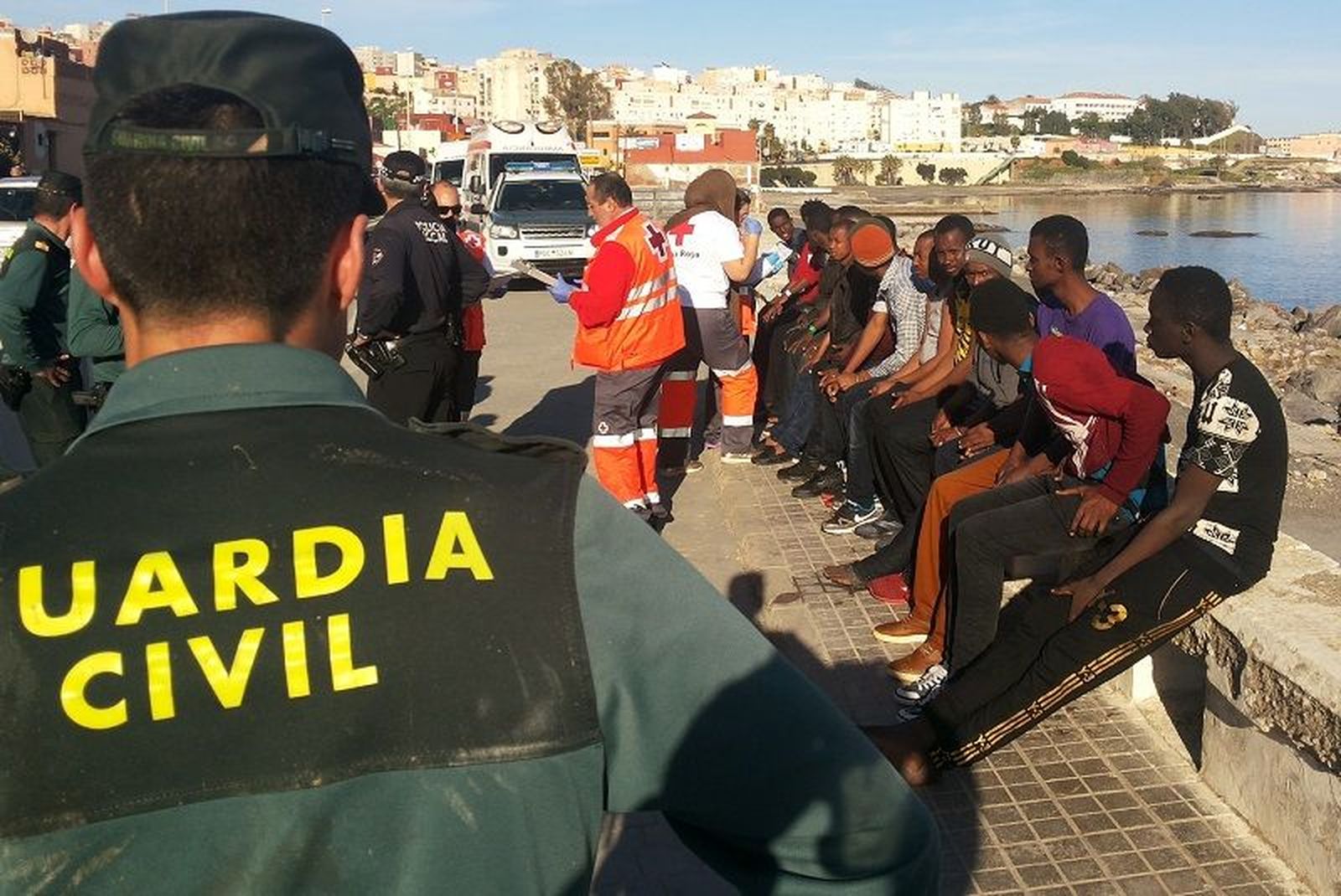 Un guardia civil observa cómo sanitarios de Cruz Roja atienden a personas migrantes recién llegadas a Ceuta