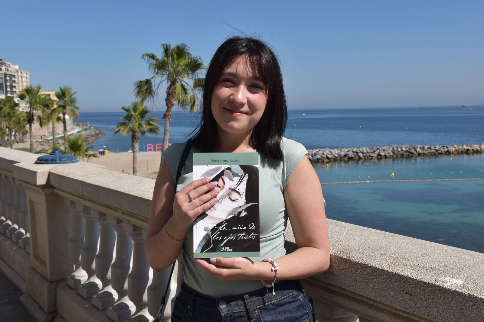 La poeta ceutí Nerea Martos posa con su primer poemario, 'La niña de los ojos tristes', frente a la playa de la Ribera