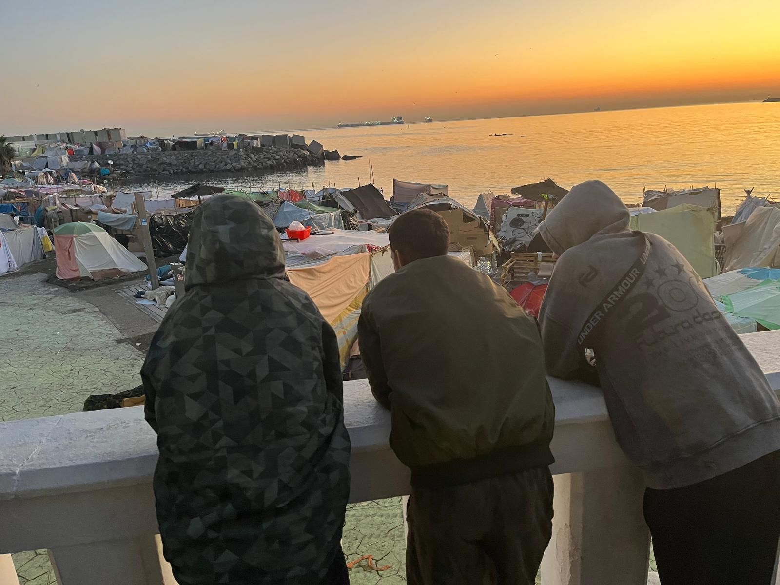 Tres chicos migrantes observan el asentamiento en playa Benítez durante el amanecer de este miércoles, cuando se esperaba que los trasladaran a instalaciones de acogida